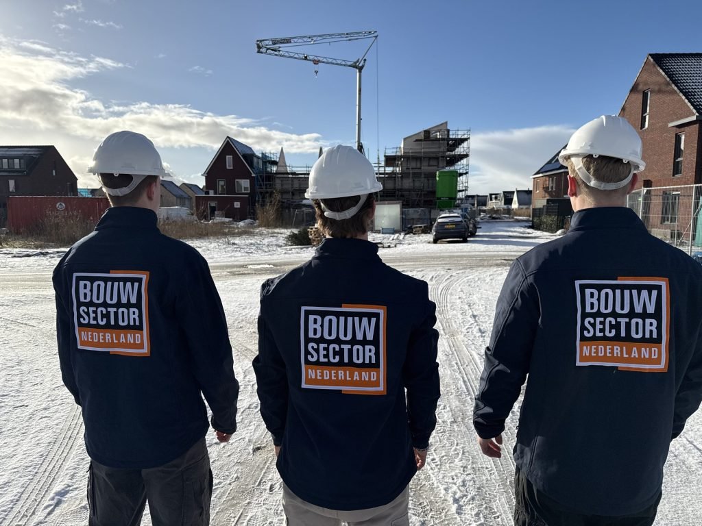 Drie teamleden van Bouwsector Nederland in donkerblauwe werkjassen met logo kijken uit over een bouwplaats